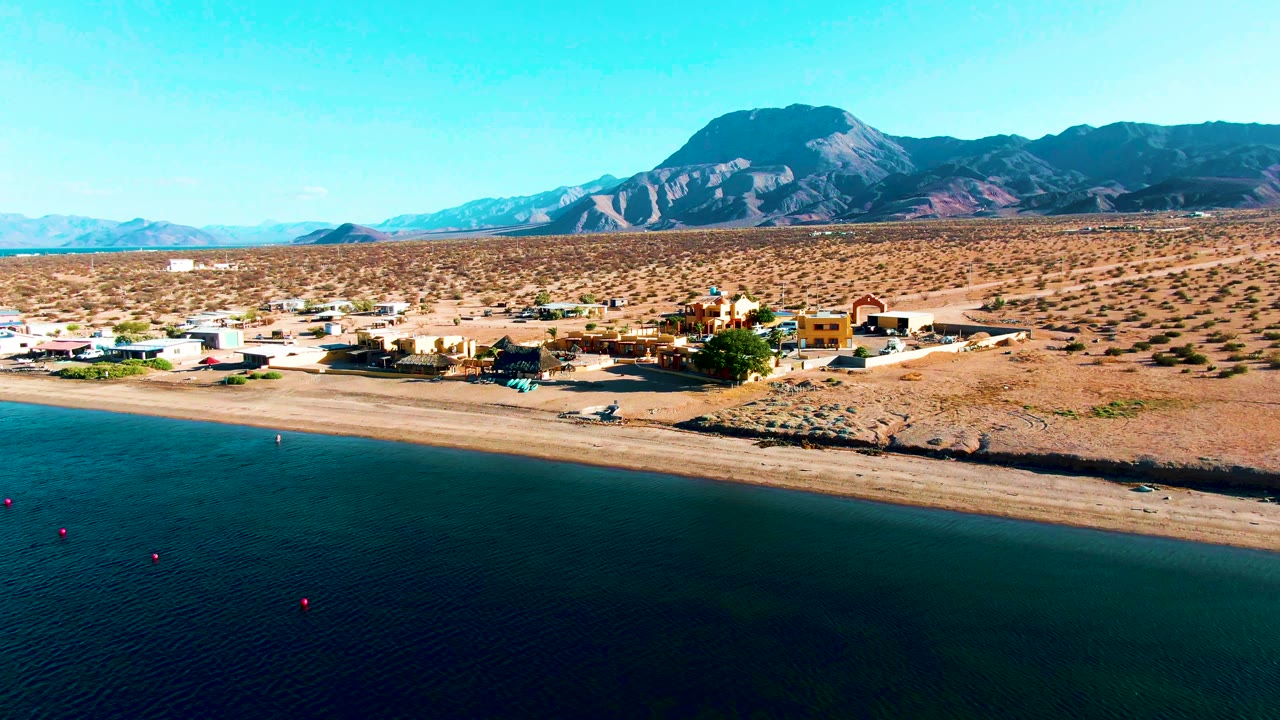 Aerial view with shoreline buildings, sandy beach, calm water, desert, and mountains.