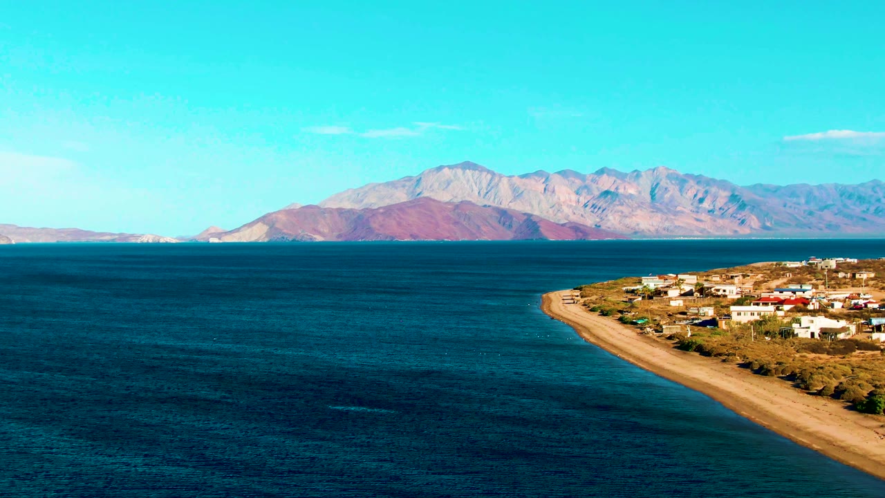 Opening frame of Bahia De Los Angeles with dark blue water in the foreground, a sandy coast and buildings at right, distant mountains across the bay, and no visible people.