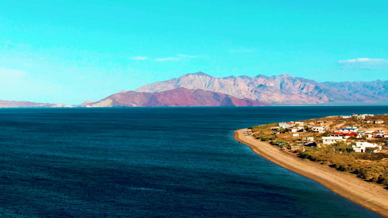 A wide Bahia De Los Angeles view shows darker ocean bands in front, the sandy beach curving past buildings at right, and mountains across the horizon, with no visible people.