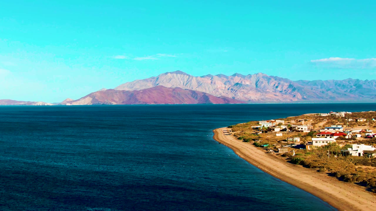 Wide aerial-looking frame of Bahia De Los Angeles with dark blue ocean in front, a sandy beach and small buildings at right, distant mountains under clear sky, and no visible people.