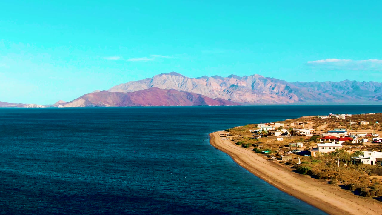 A wide 4K frame of Bahia De Los Angeles showing the beach running along the right edge, scattered shoreline buildings, deep blue water, distant mountains, and no visible people.