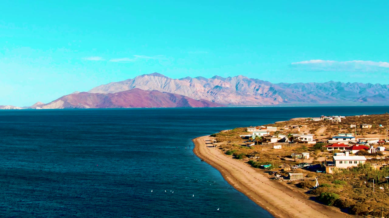 Opening frame of Bahia De Los Angeles with a sandy coastal road, shoreline buildings, blue ocean, distant mountains, clear sky, and no visible people.