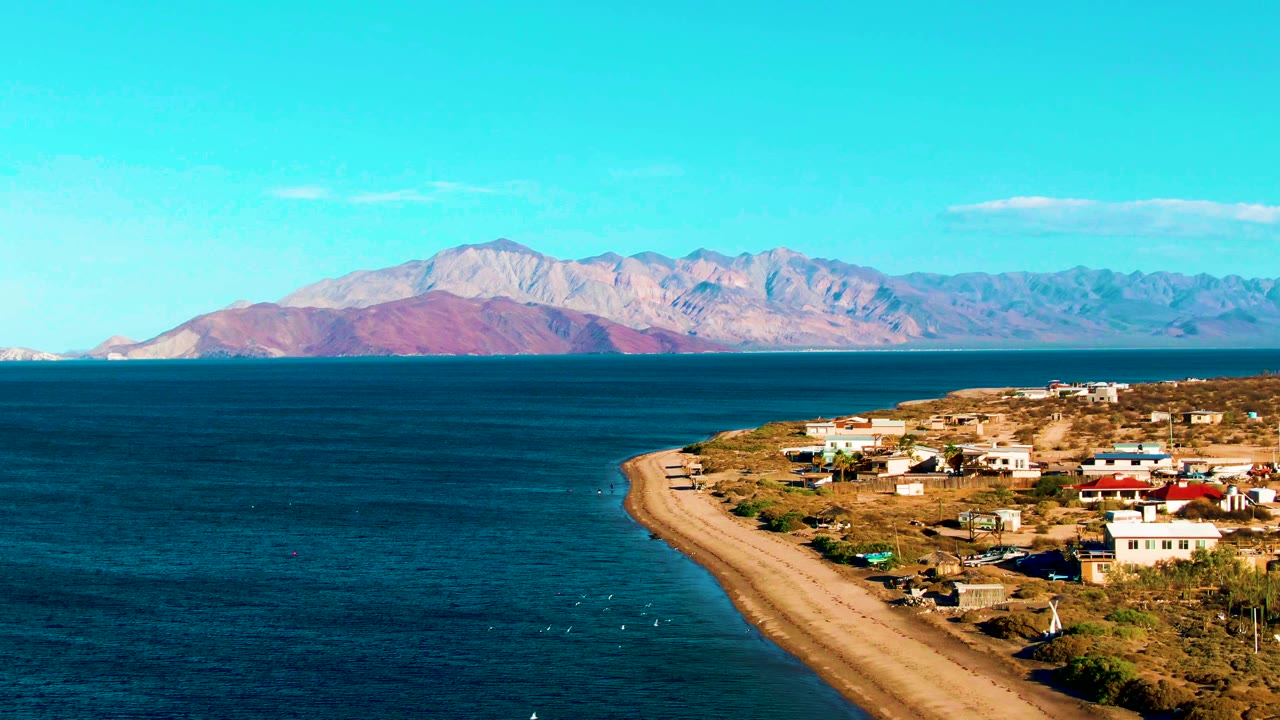One second into the clip, Bahia De Los Angeles is seen from above with dark blue water, a curved sandy shore road, scattered buildings, distant mountains, and no visible people.