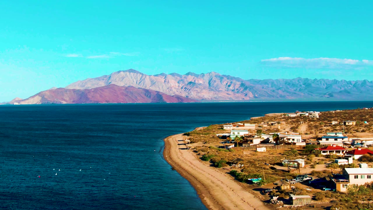 Opening frame of Bahia De Los Angeles with a curved sandy shore, blue ocean, dry coastal settlement, distant mountains, clear sky, and no visible people.