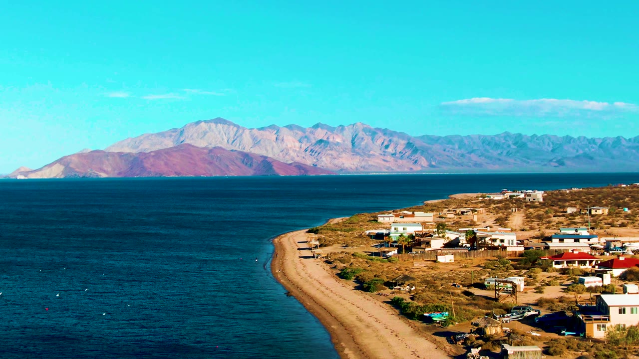 Bahia De Los Angeles coast at 0:01, showing the beach line closer to center, deep blue water to the left, scattered buildings to the right, mountains beyond, and no visible people.