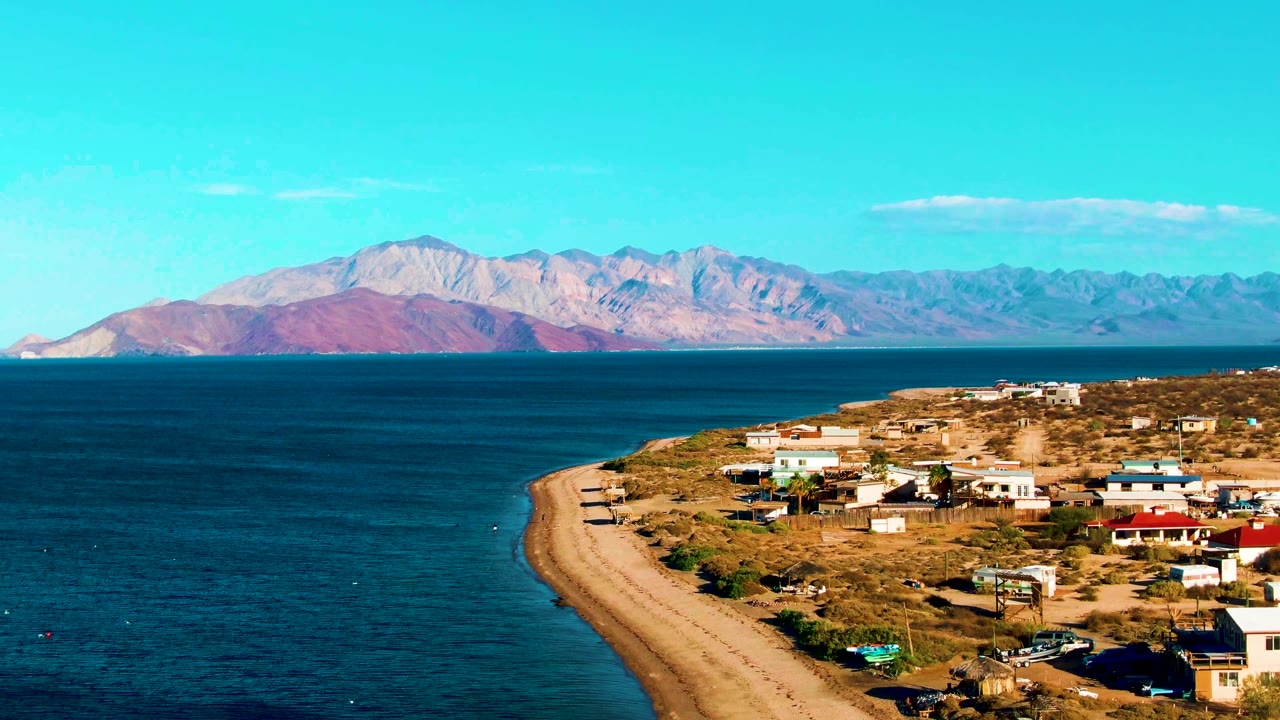 Bahia De Los Angeles shoreline at 0:02 with the settlement spread across the right side, the sandy beach tracing the bay, open water on the left, a clear sky, and no visible people.