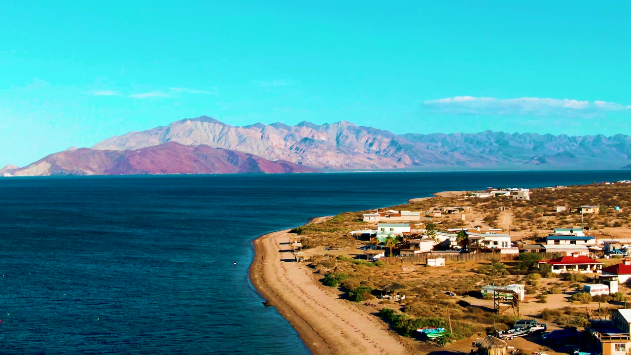 Blue ocean and sandy beach beside low buildings with desert hills behind