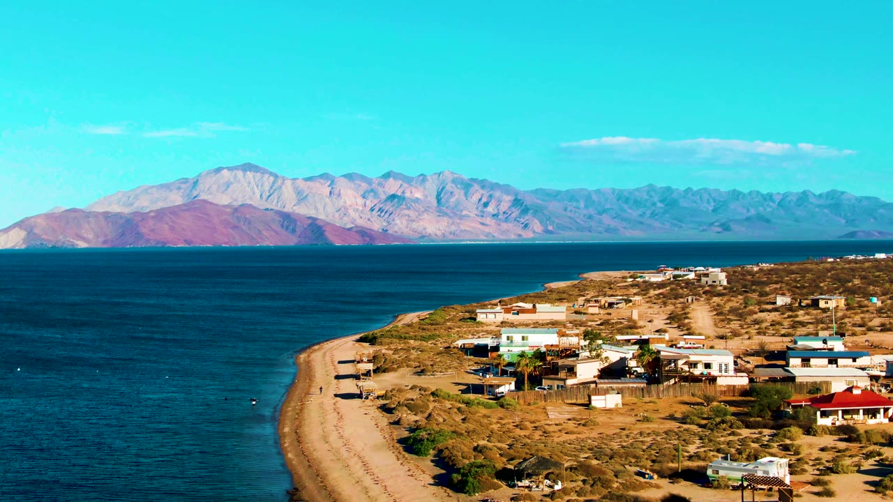Opening aerial frame over Bahia De Los Angeles, Mexico, with a curved sandy shore, blue ocean, scattered coastal buildings, dry terrain, and mountains across the water.