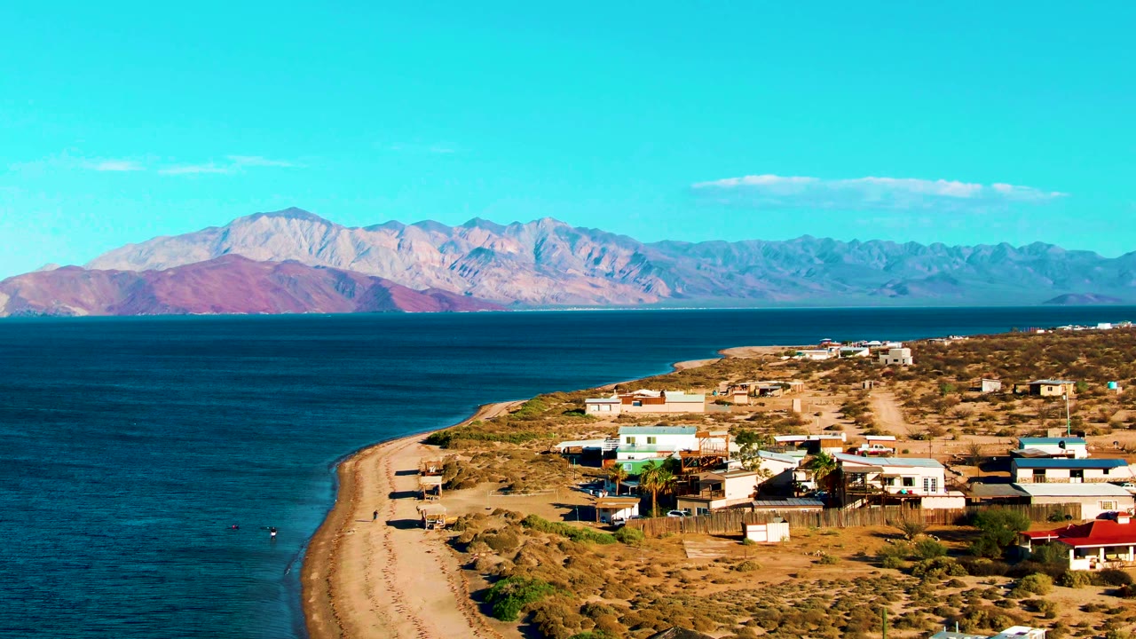 At 0:02, Bahia De Los Angeles appears as a broad coastal aerial with the beach running up the left side, village buildings at right, dark water in midframe, and mountains on the horizon.