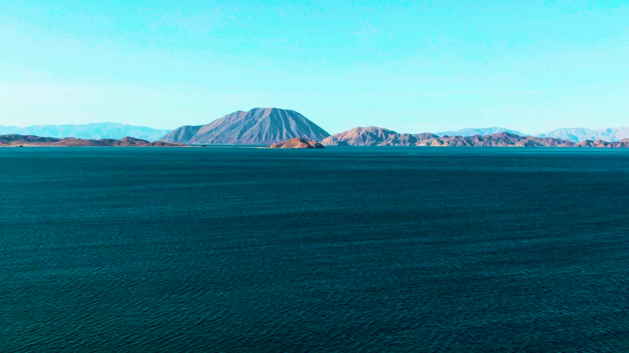 At 0:01, Bahia De Los Angeles appears as a broad expanse of dark blue water with low mountains and a rounded landform across the horizon, with no visible people.