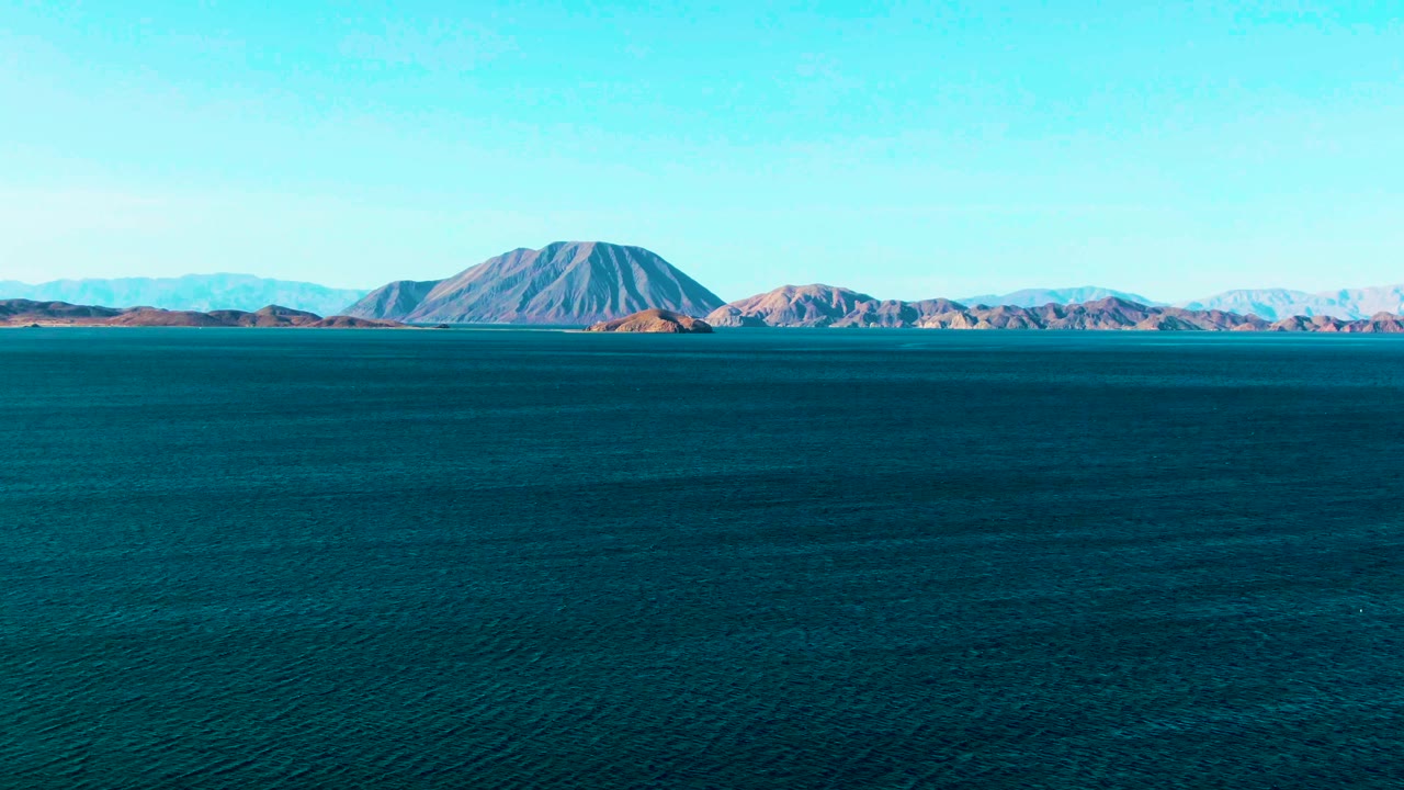 At 0:02, the frame holds on rippled blue sea and a distant rounded mountain backed by lower ridges in Bahia De Los Angeles, with clear sky and no visible people.