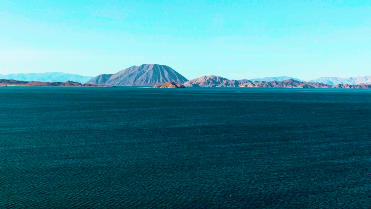 At 0:03, dark blue water fills the lower frame while the Bahia De Los Angeles mountain landmass stretches across the horizon beneath a cloudless-looking sky, with no visible people.
