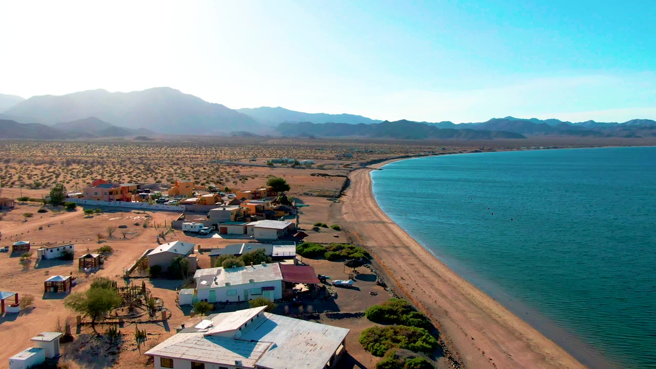 Aerial view over Bahia De Los Angeles with low beach houses in the foreground, a sandy shoreline curving beside turquoise water, desert scrub, distant mountains, and no visible people.