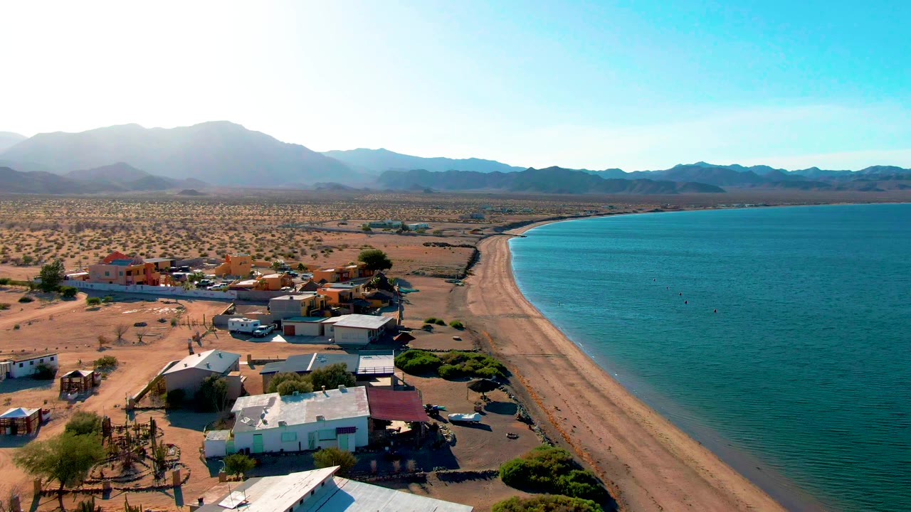 Drone view of curved sandy beach beside a small desert settlement