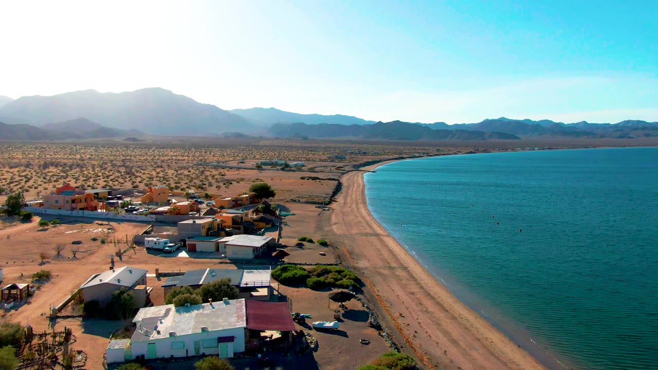Elevated frame over Bahia De Los Angeles with rooftops and a few vehicles along the lower edge, the sandy beach running into the distance, calm turquoise water on the right, desert plain on the left, and no visible people.