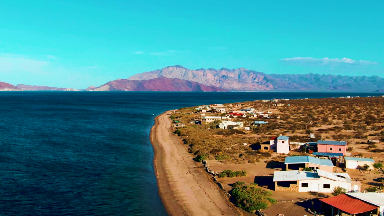 Aerial frame over Bahia De Los Angeles showing blue ocean at left, a sandy shoreline, low coastal buildings on dry scrubland, distant mountains, and small people visible near the beach.