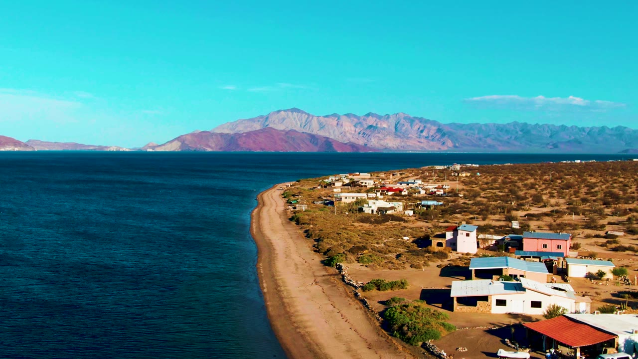 Drone view of shoreline curving past village buildings toward distant mountains.