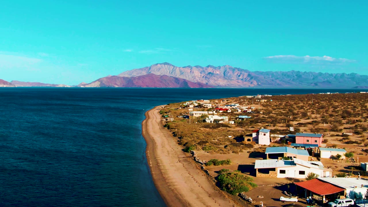 Wide drone frame over Bahia De Los Angeles showing low waterfront buildings, a red-roofed structure in the foreground, sandy beach, blue bay water, distant mountains, and tiny people near the shore.