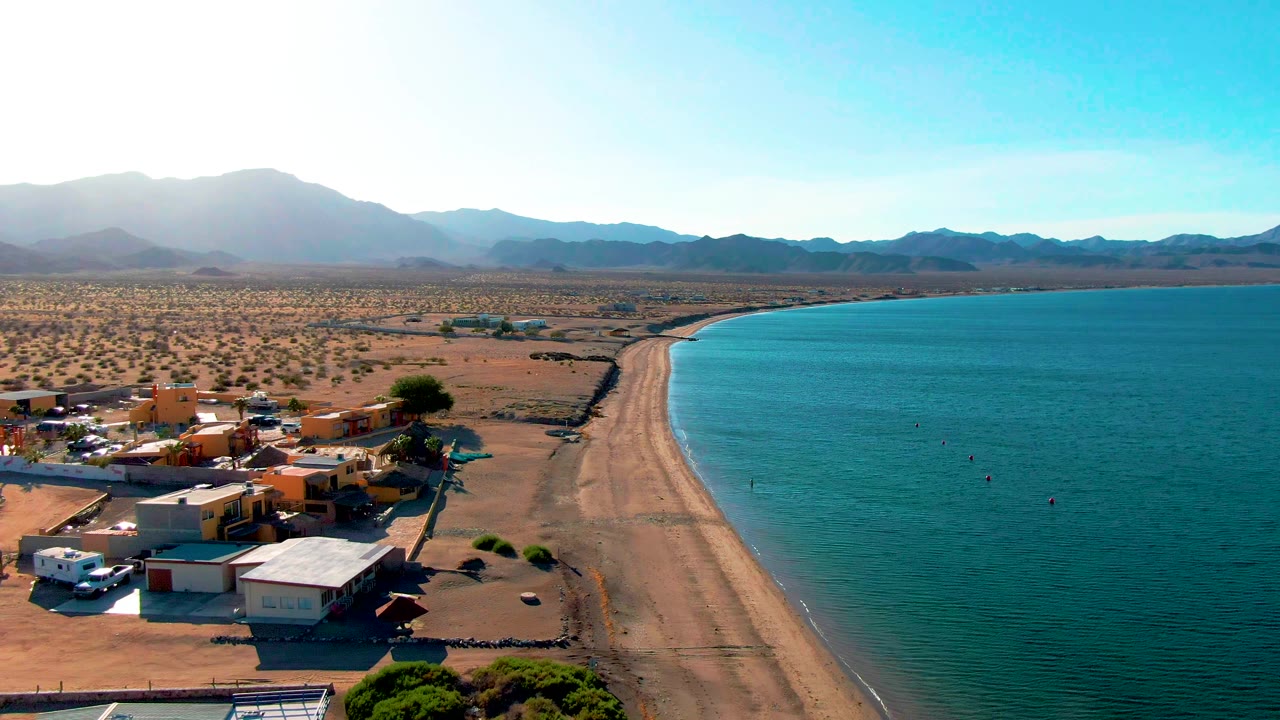 Opening aerial frame over Bahia De Los Angeles shows low shoreline buildings and parked vehicles at left, a sandy beach curving beside blue water, desert flats, distant mountains, and no visible people.
