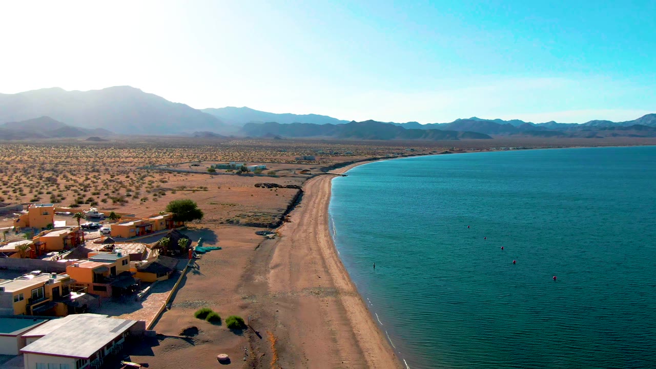 At 0:02, the Bahia De Los Angeles shoreline sits under bright light with orange and white buildings near the lower left, a long beach, open blue water on the right, desert flats, mountains, and no visible people.