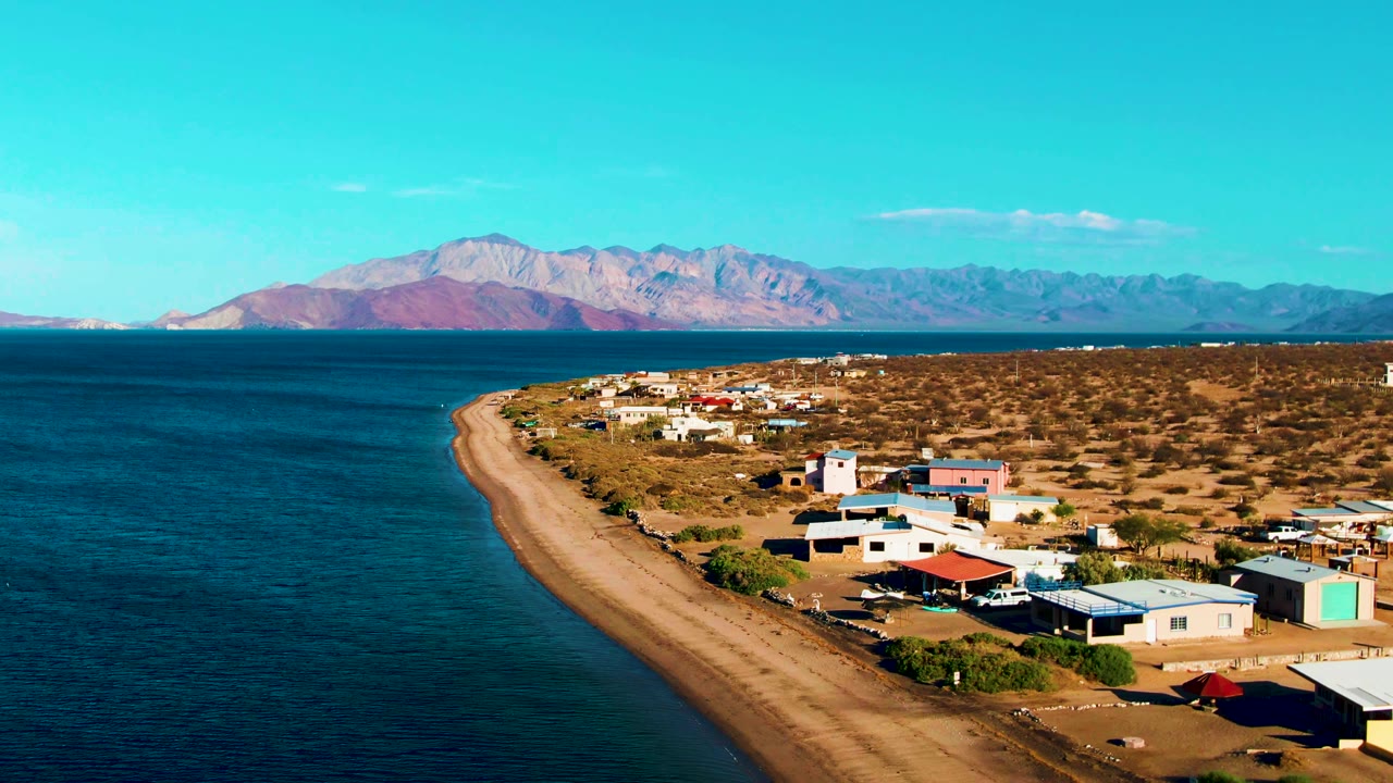 Opening frame of Bahia De Los Angeles with blue ocean at left, a sandy curved shoreline, scattered desert buildings at right, distant mountains, clear sky, and no visible people.