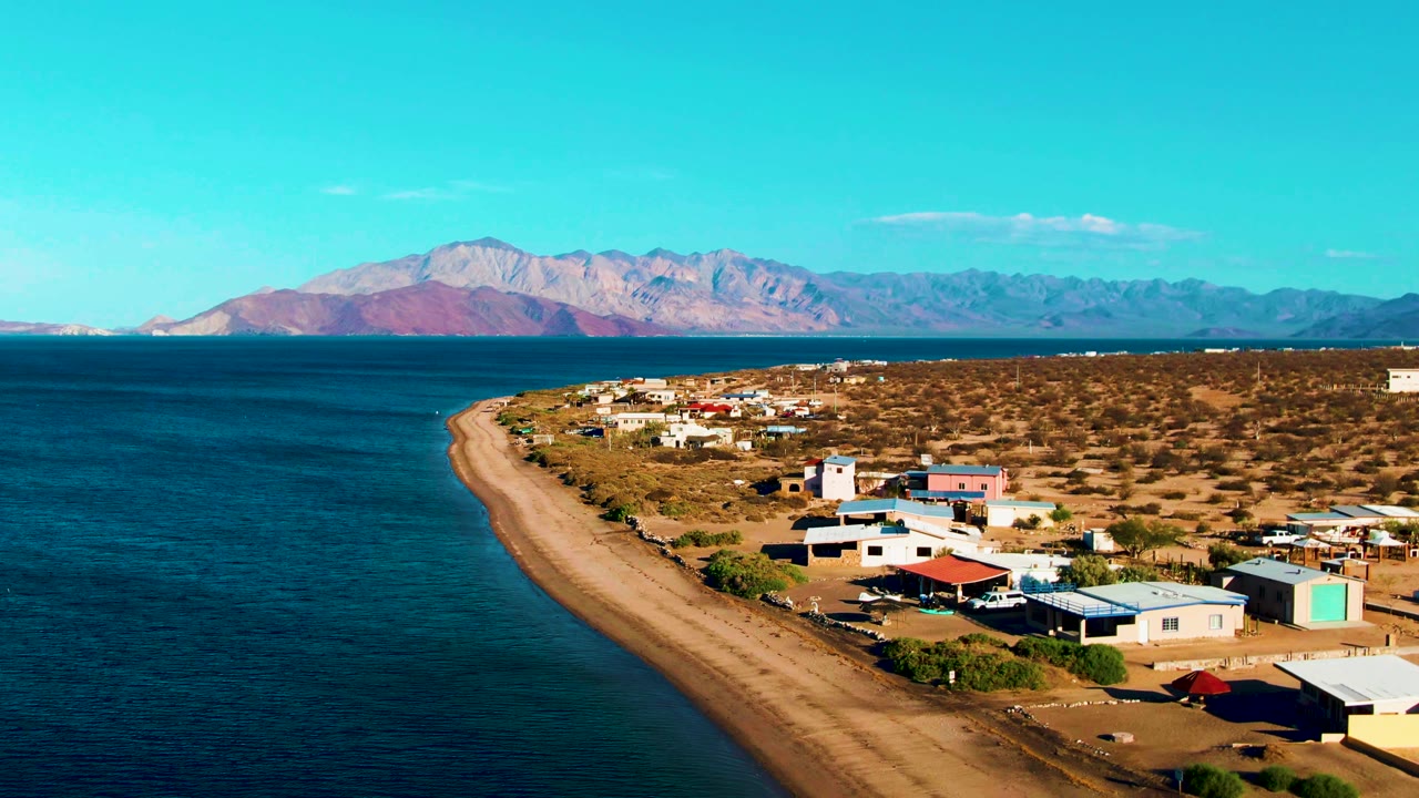 At 0:01, Bahia De Los Angeles appears from above with the ocean stretching along the left edge, tan beach curving inward, low buildings in the foreground, mountains beyond the bay, and no visible people.