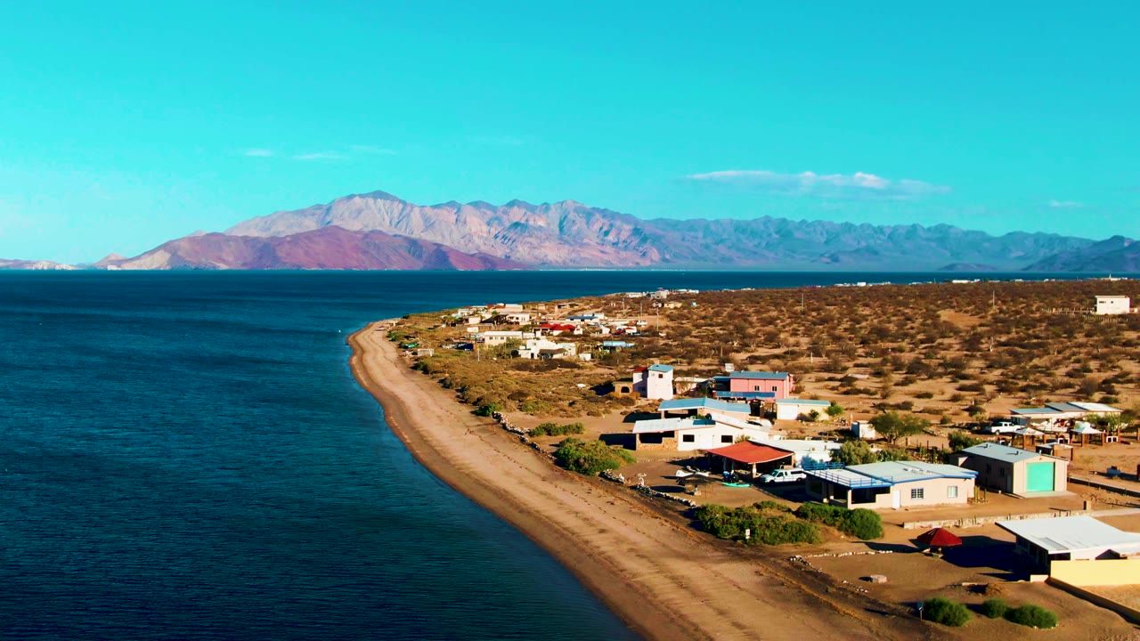 At 0:02, the aerial view keeps the sandy Bahia De Los Angeles shoreline in the center, dark blue water to the left, small buildings and open desert to the right, mountain ridges in back, and no visible people.
