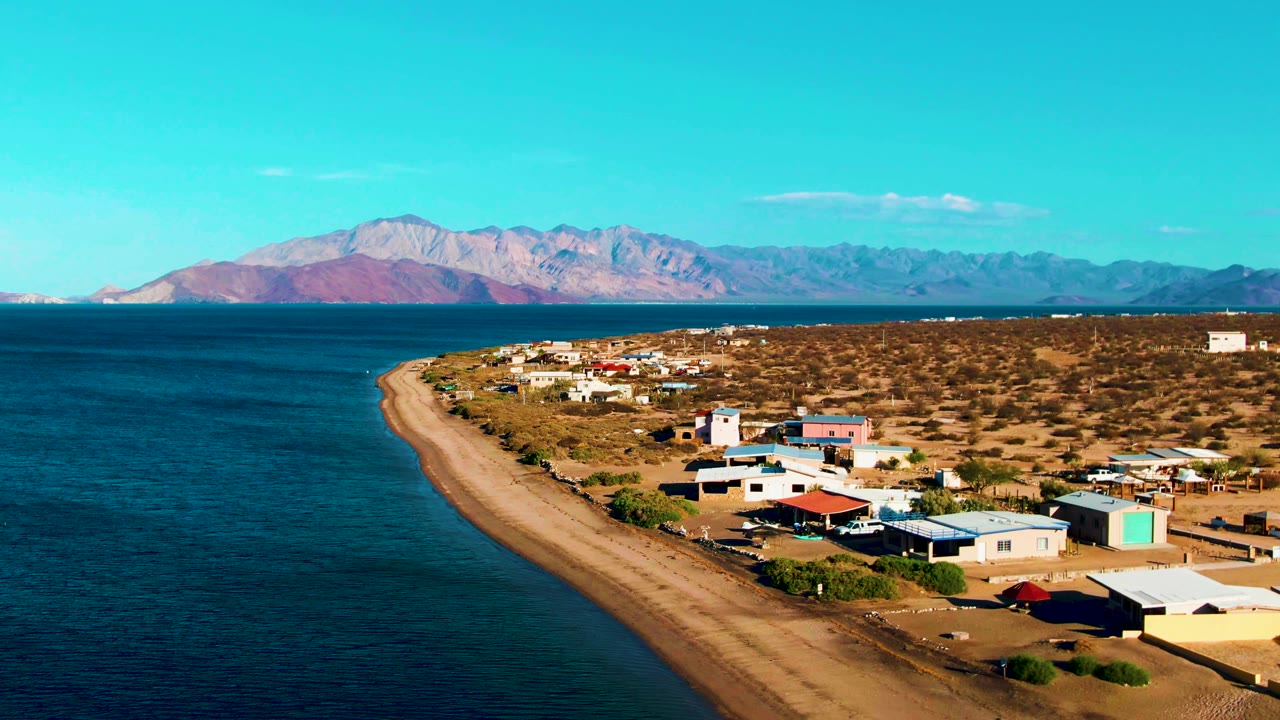 High coastal view over blue water, tan beach, desert settlement, and mountain horizon.