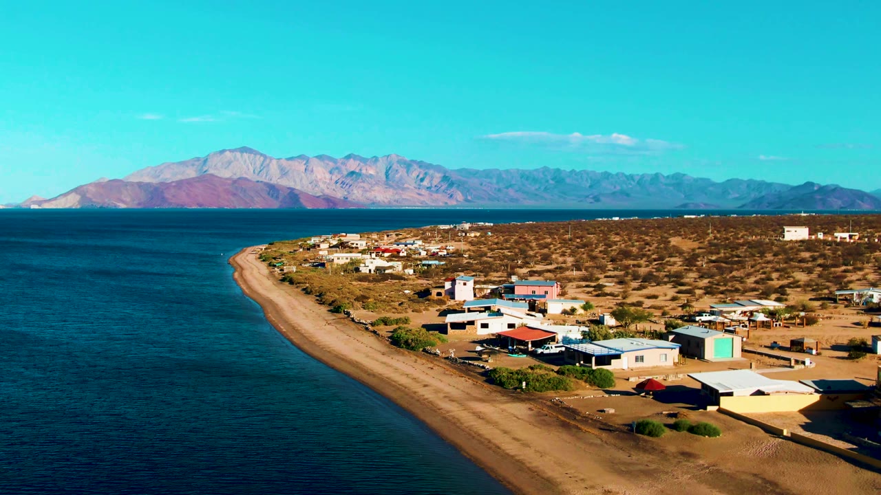 First frame of Bahia De Los Angeles showing blue ocean beside a curving sandy shoreline, scattered village buildings, desert scrub, distant mountains, and no visible people.