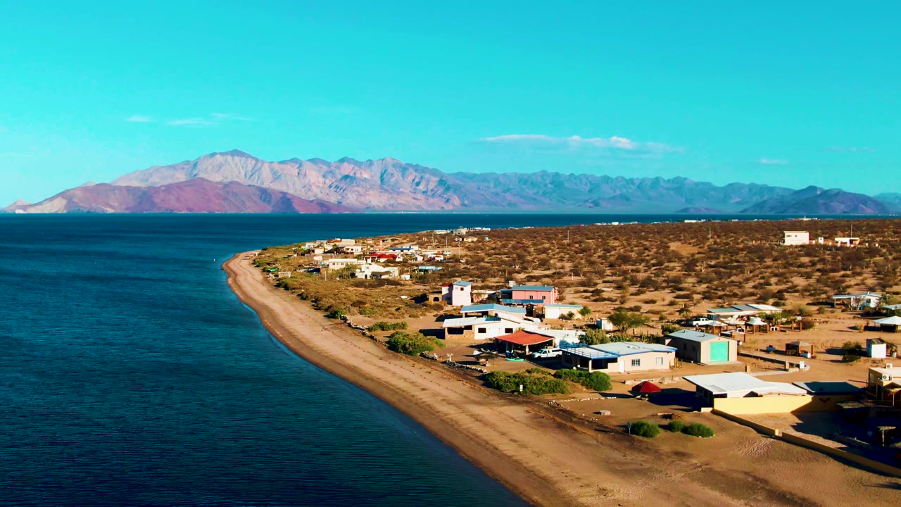 At 0:01, Bahia De Los Angeles appears from a high oblique angle with dark blue water, a pale beach curve, low coastal buildings, arid ground, and no visible people.