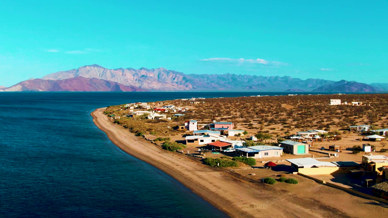 At 0:02, the frame shows the Bahia De Los Angeles shoreline running diagonally through the scene, with ocean to the left, buildings to the right, mountains beyond, and no visible people.