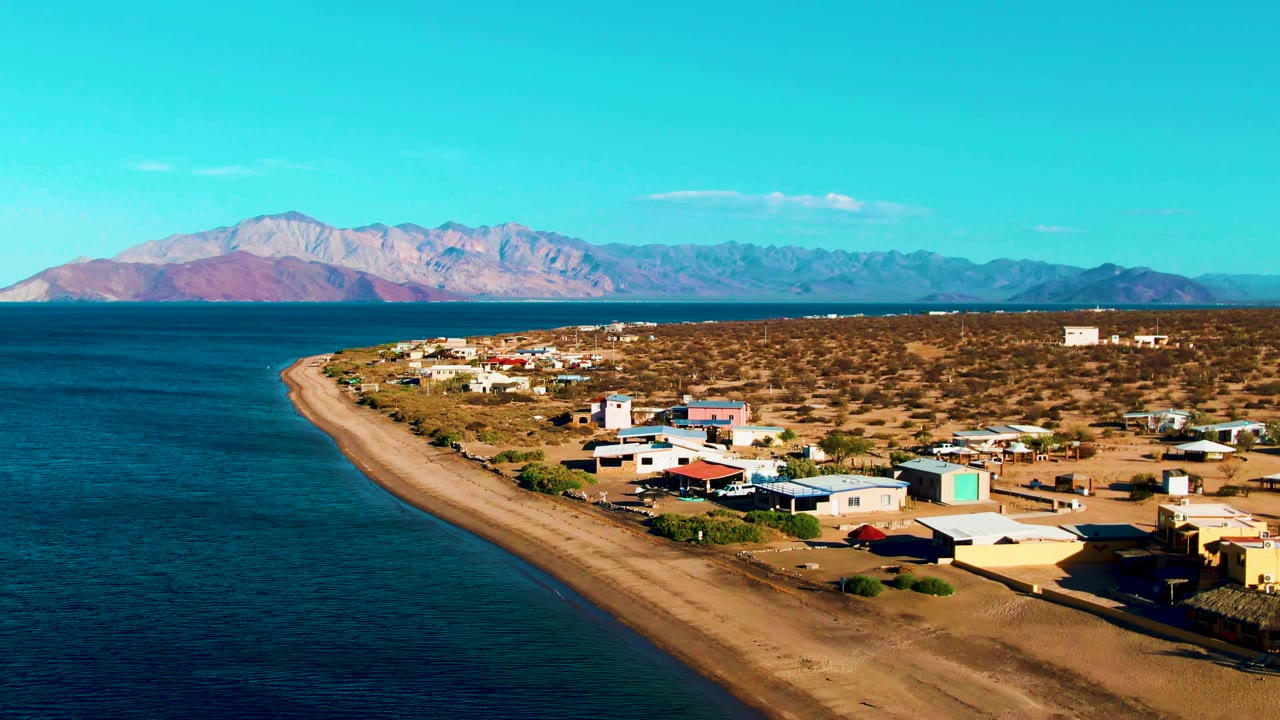 Aerial coastline view with calm water, sandy beach, small structures, and mountain backdrop.