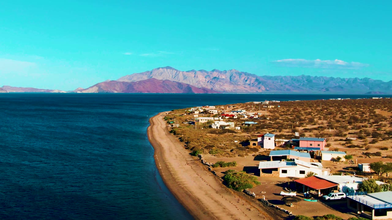 Opening aerial frame over Bahia De Los Angeles shows deep blue ocean at left, a curving sandy beach, scattered beachside buildings, dry terrain, distant mountains, and no visible people.
