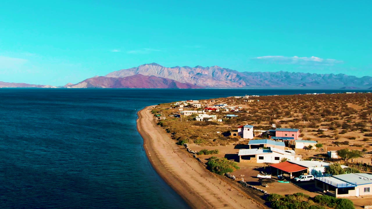 At 0:02, Bahia De Los Angeles appears from an elevated angle with more right-side beach houses in view, the ocean occupying the left half, and no visible people.