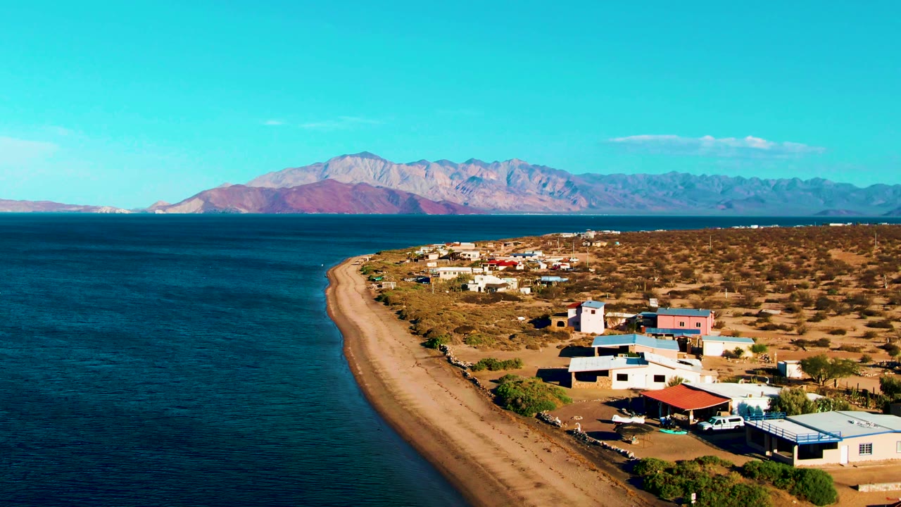 Blue water, sandy coastal road, scattered buildings, and mountain horizon.