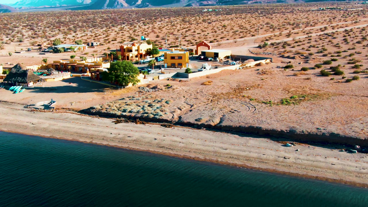Aerial view of teal water, sandy shore, desert buildings, and distant mountains.