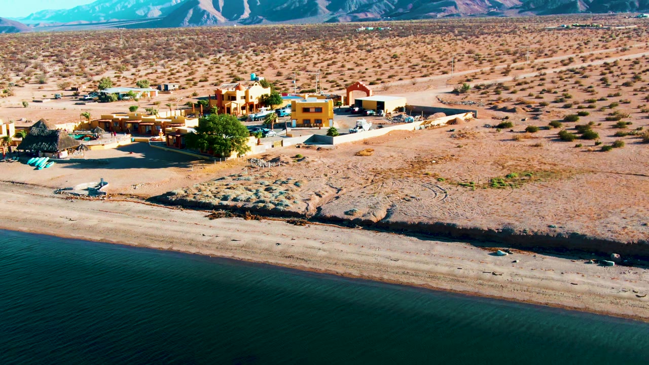 Aerial view in Bahia De Los Angeles with the village shifted near center frame, calm water below, sandy beach, scrubby desert plain, mountains, and no visible people.