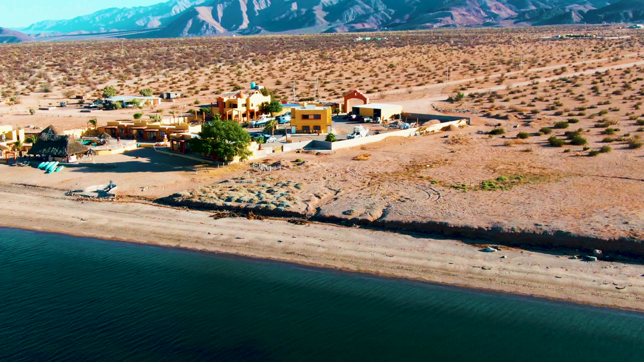 Wide aerial frame from Bahia De Los Angeles showing turquoise water, a pale beach strip, colorful low buildings, broad desert flats, distant mountains, and no visible people.