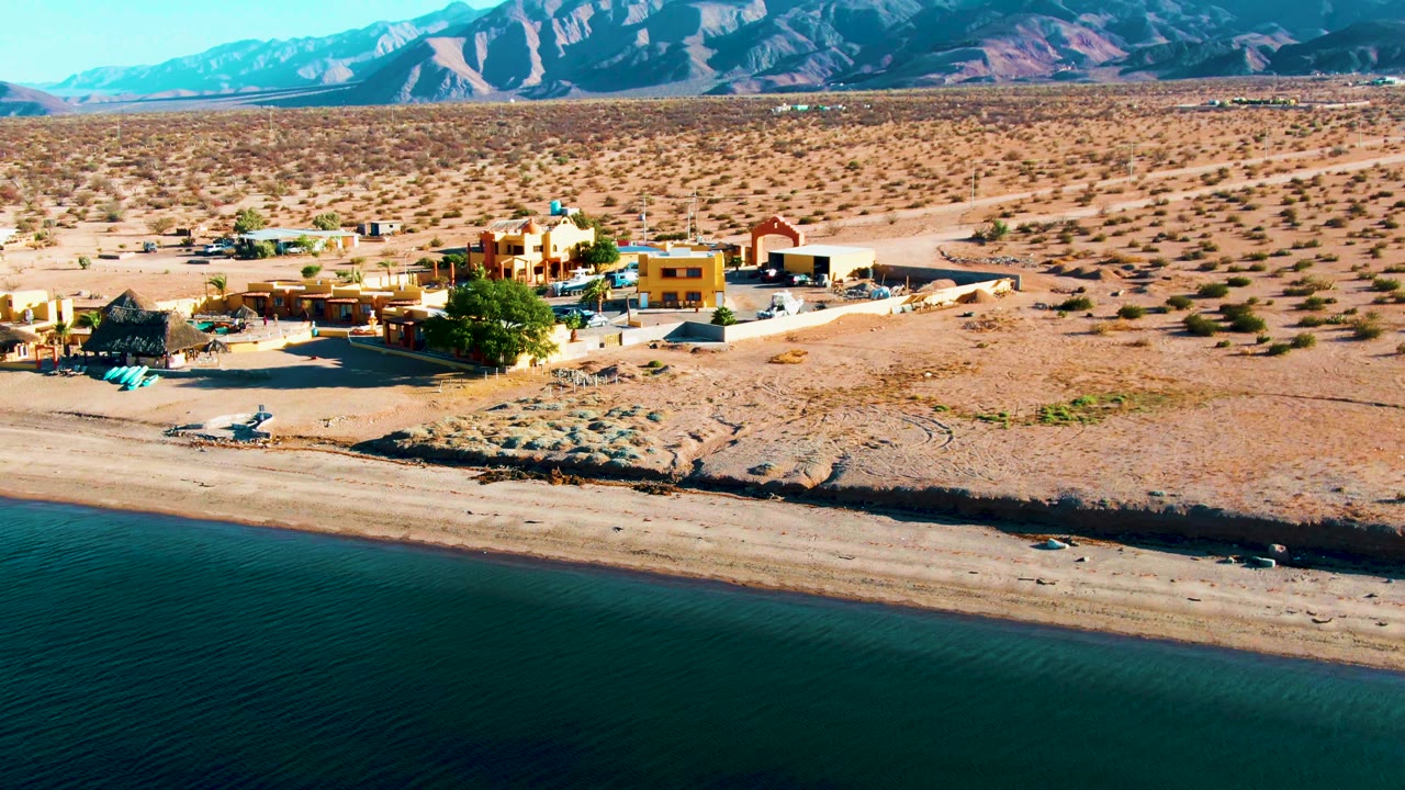 Aerial view over Bahia De Los Angeles with dark teal water at the bottom, a sandy shoreline, village buildings to the left of center, desert scrub, mountains, and no visible people.