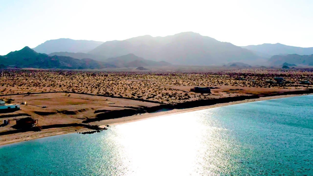 Opening aerial frame over Bahia De Los Angeles with bright turquoise water below, a sandy shoreline, arid desert plain, hazy mountains, and no visible people.