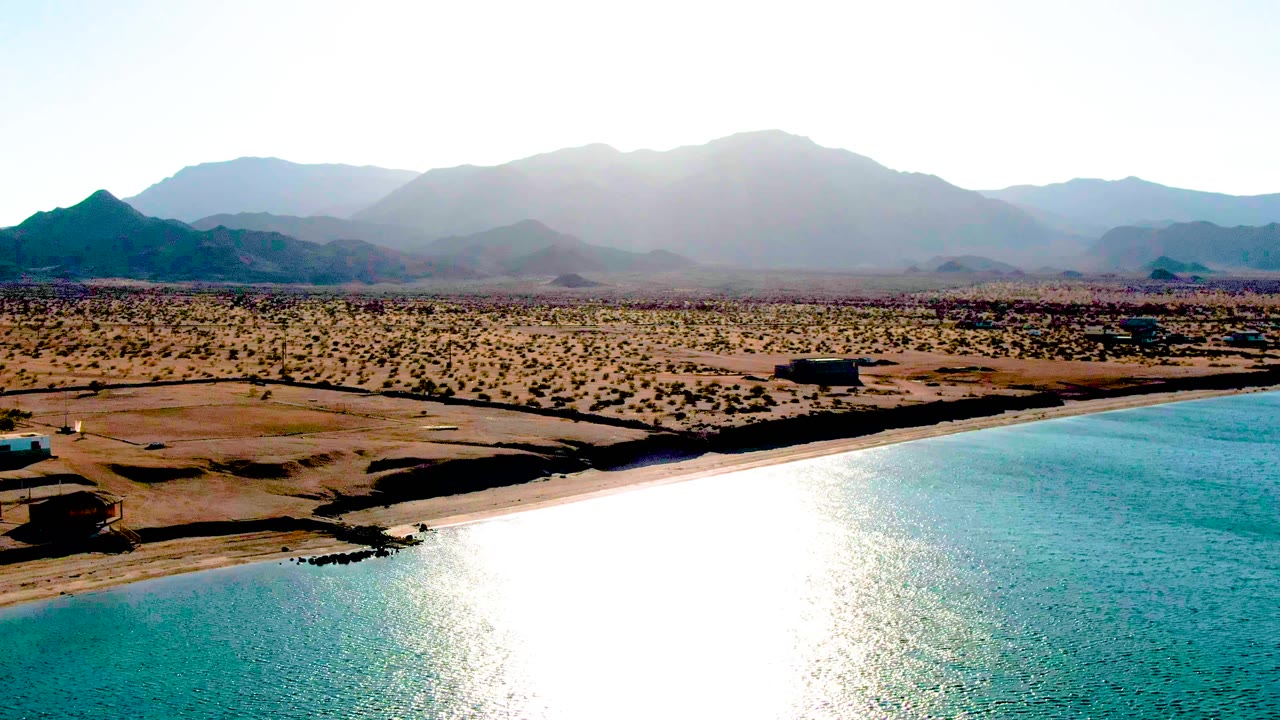 Bahia De Los Angeles coastline seen from above with a strong white sun reflection on the ocean, tan shore, open desert terrain, layered mountains, and no visible people.