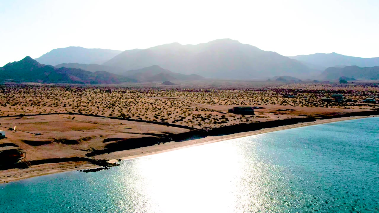 Aerial frame over Bahia De Los Angeles showing the beach curving beside turquoise water, sparse desert vegetation, low structures or dark objects inland, hazy mountain ridges, and no visible people.