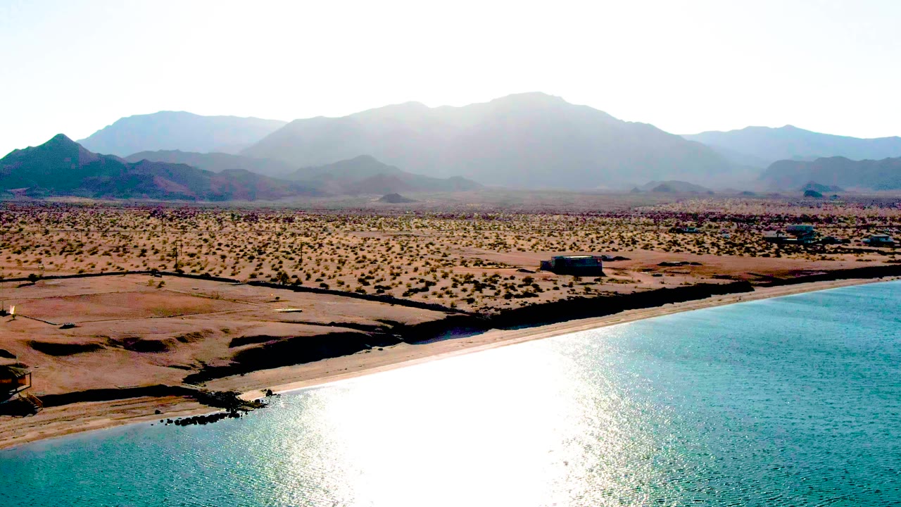 Mid-clip aerial view of Bahia De Los Angeles with intense light reflected on the water, a narrow sandy shore, open brown desert, distant mountain silhouettes, and no visible people.
