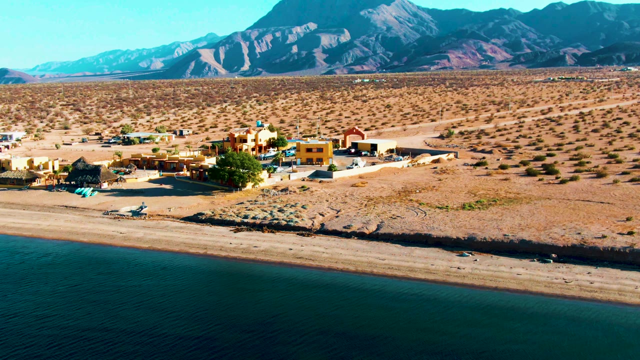 At 0:00, Bahia De Los Angeles appears from above with dark calm water at the bottom, a sandy beach, a cluster of small buildings, arid scrubland, rugged mountains, and no visible people.