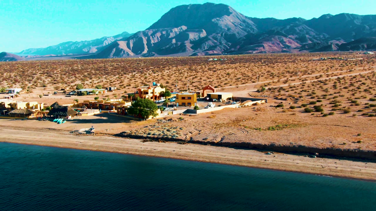 At 0:02, the Bahia De Los Angeles beach and small building cluster sit between calm blue water and a dry desert plain, with the mountain ridge filling the background and no visible people.