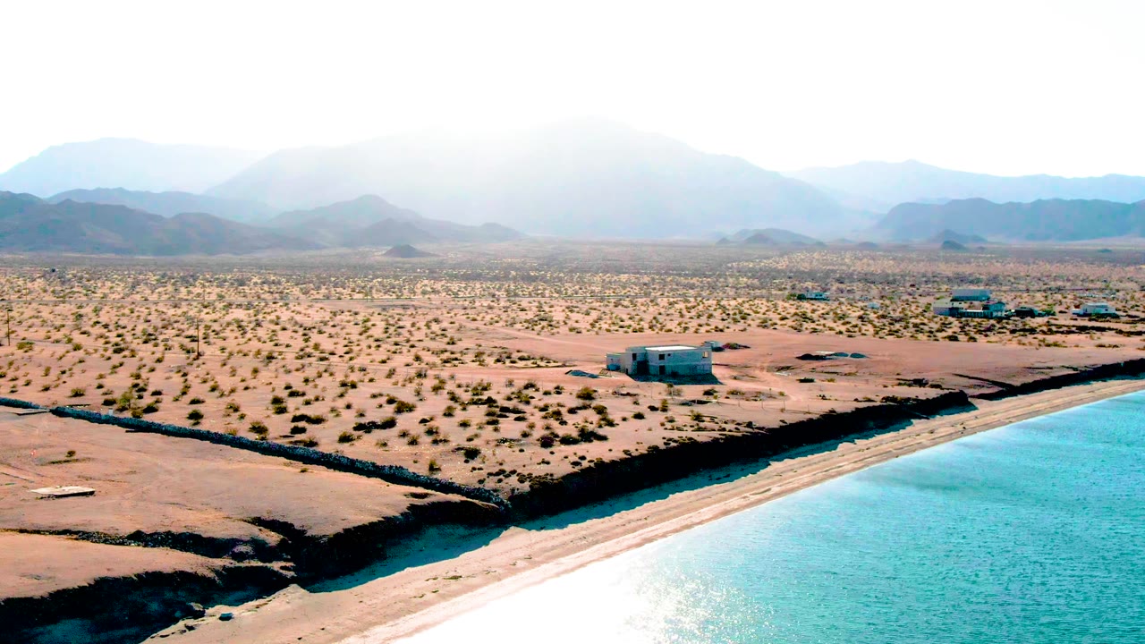 Aerial frame over Bahia De Los Angeles showing turquoise water at the lower right, sandy desert scrub, small low structures, distant hazy mountains, and no visible people.