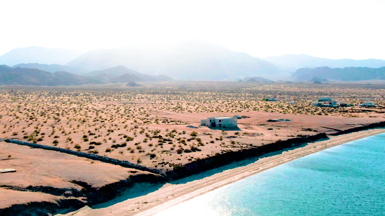 Drone-style aerial frame in Bahia De Los Angeles with the water edge filling more of the lower right, a dark canal or shoreline border, scrubby desert, low buildings, mountains, and no visible people.
