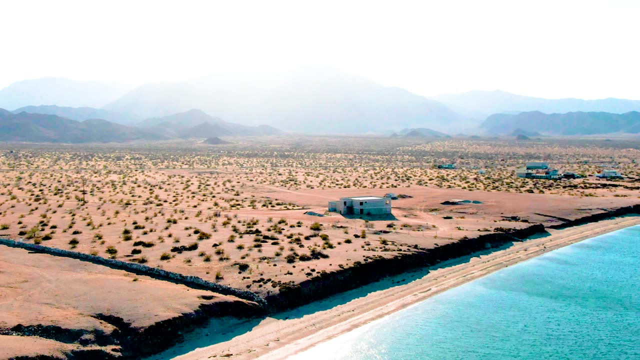 Wide aerial frame over Bahia De Los Angeles with a bright blue water edge in the foreground corner, sandy scrubland across the center, scattered small structures, layered mountains, and no visible people.