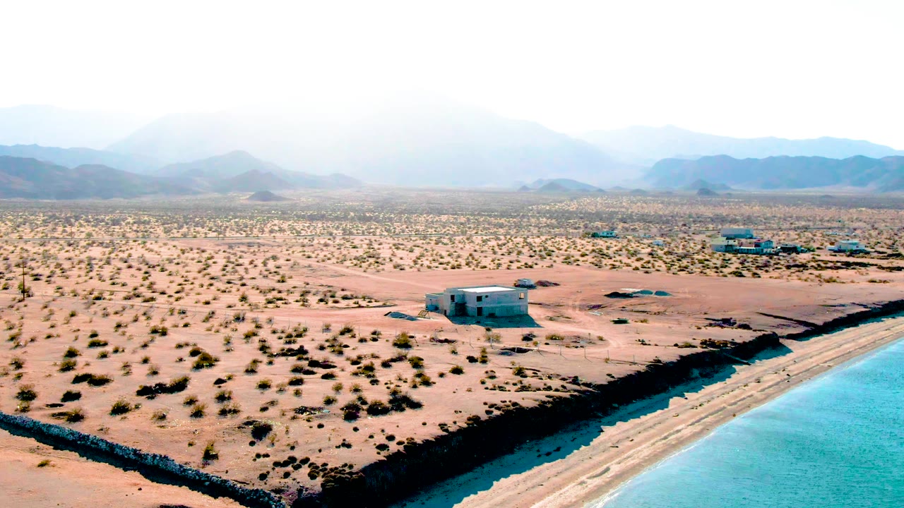 Aerial frame over Bahia De Los Angeles showing turquoise reservoir water at lower right, sandy desert with sparse shrubs, small structures in the midground, hazy mountains beyond, and no visible people.