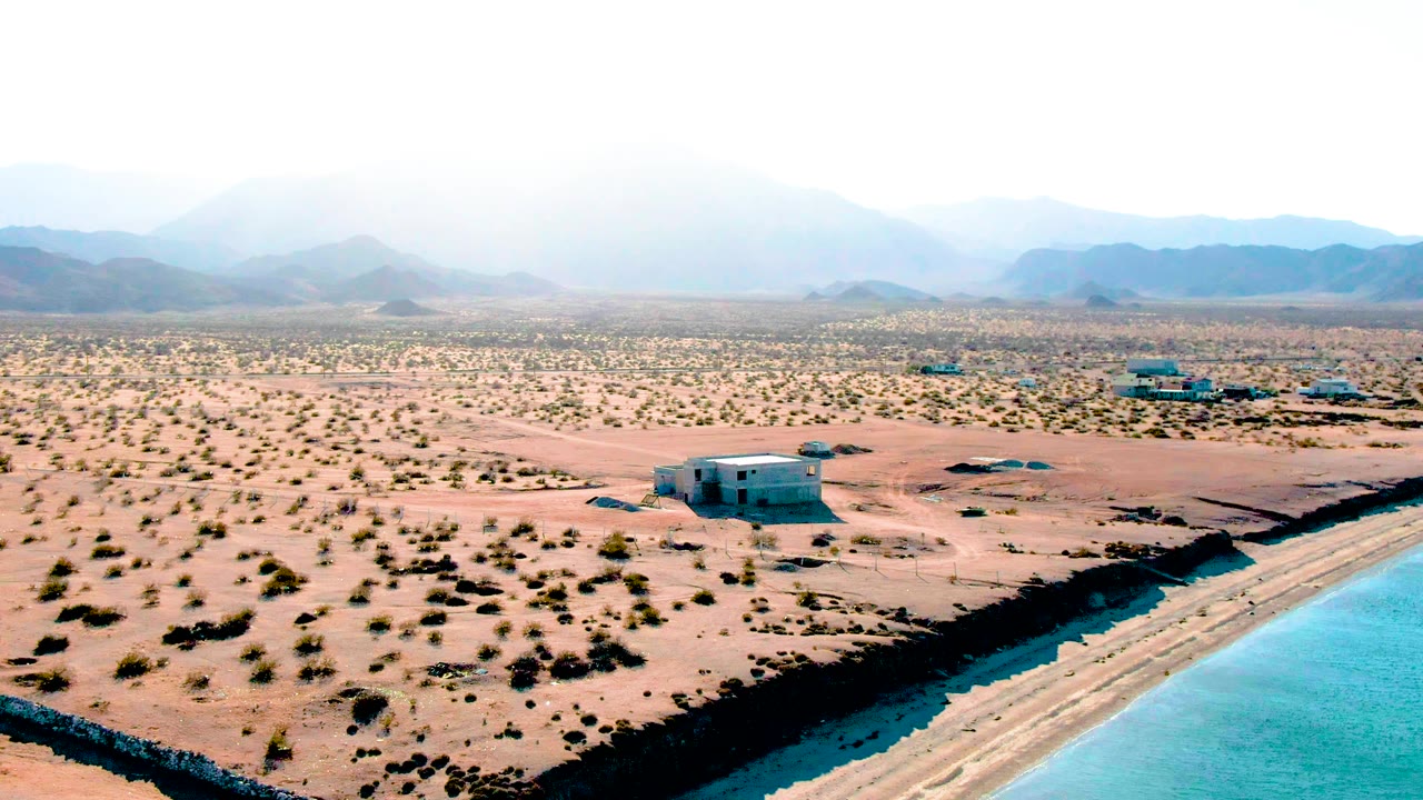 Wide aerial view in Bahia De Los Angeles with the reservoir edge cutting along the lower right, dry sandy ground and scrub across the center, low buildings or trailers in the distance, and no visible people.